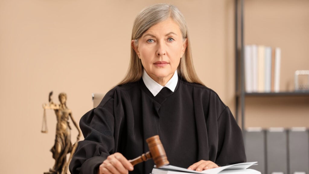 A judge in a black robe holds a gavel, sitting at a desk with an open book and a Lady Justice statue.