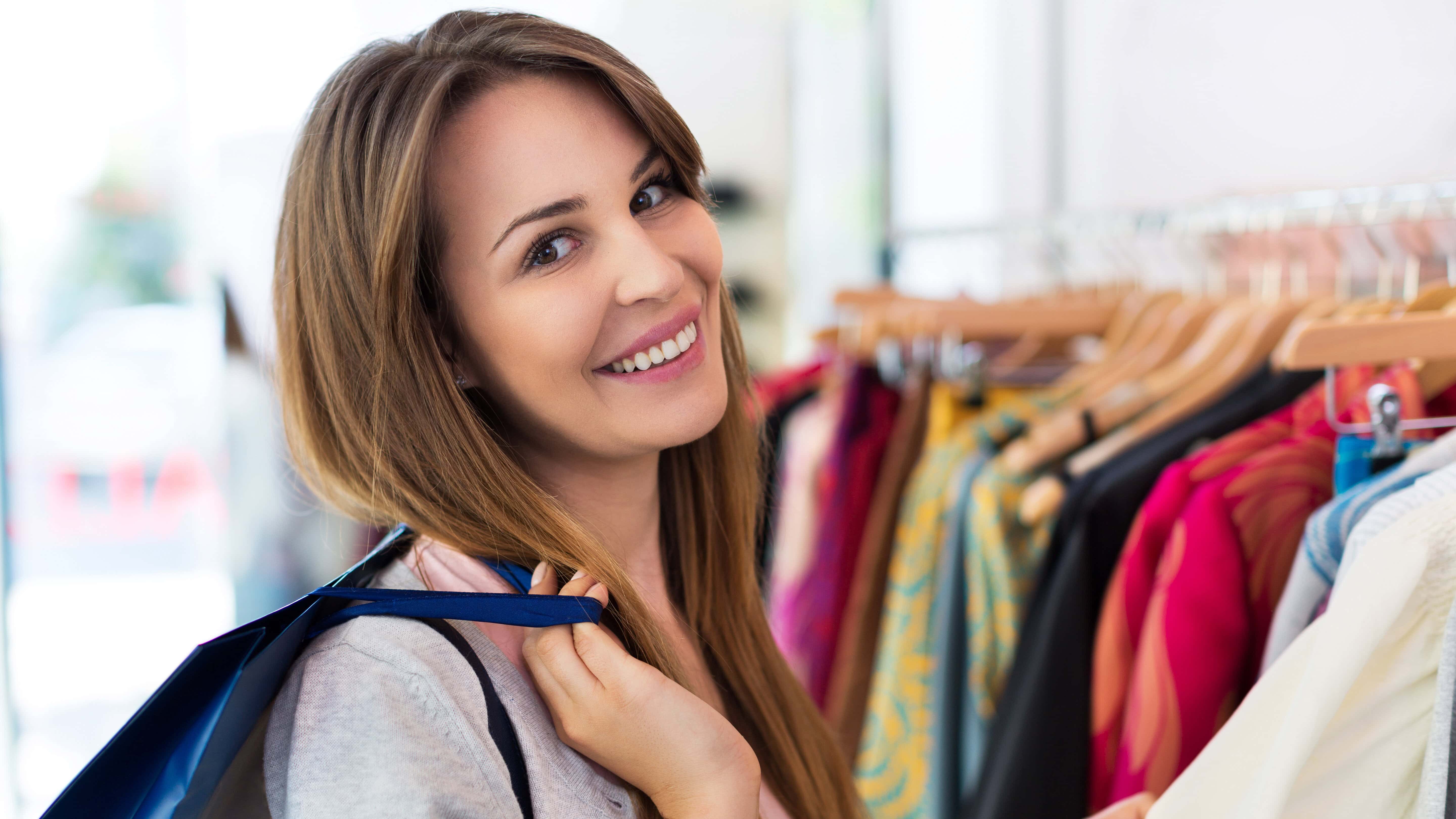 Woman smiling and holding a shopping bag over her shoulder while standing near a clothing rack in a store.