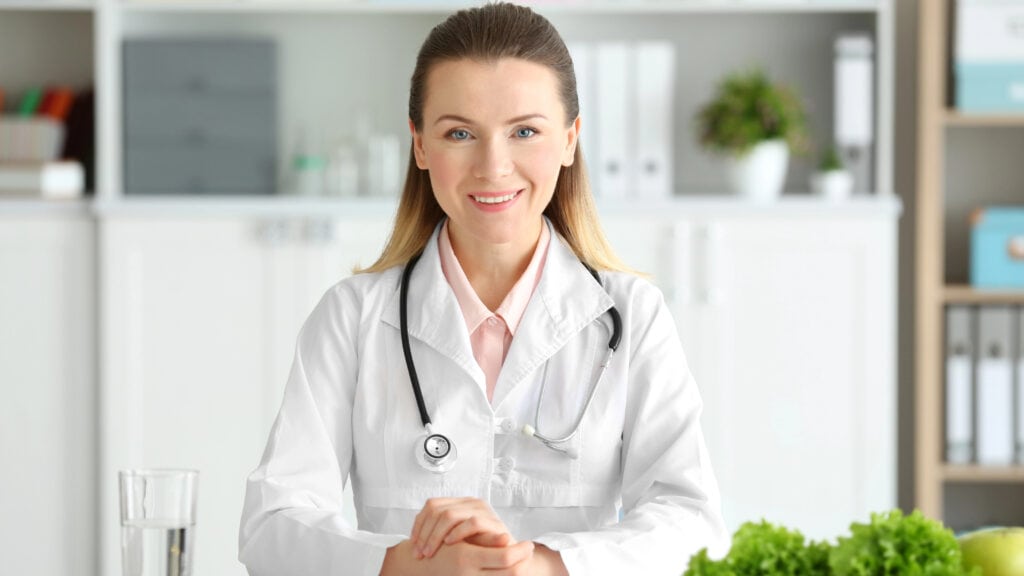 A female doctor with a stethoscope sits at a desk surrounded by fresh vegetables in a medical office.