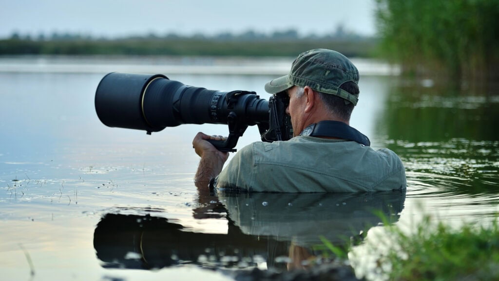 A man in water, wearing a cap and holding a large camera with a telephoto lens, is focused on capturing a distant subject.