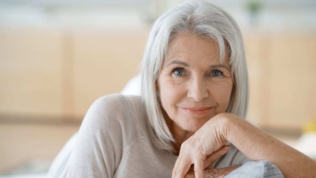 An elderly woman with white hair and a light-colored top sits and smiles while resting her hands on her chin.