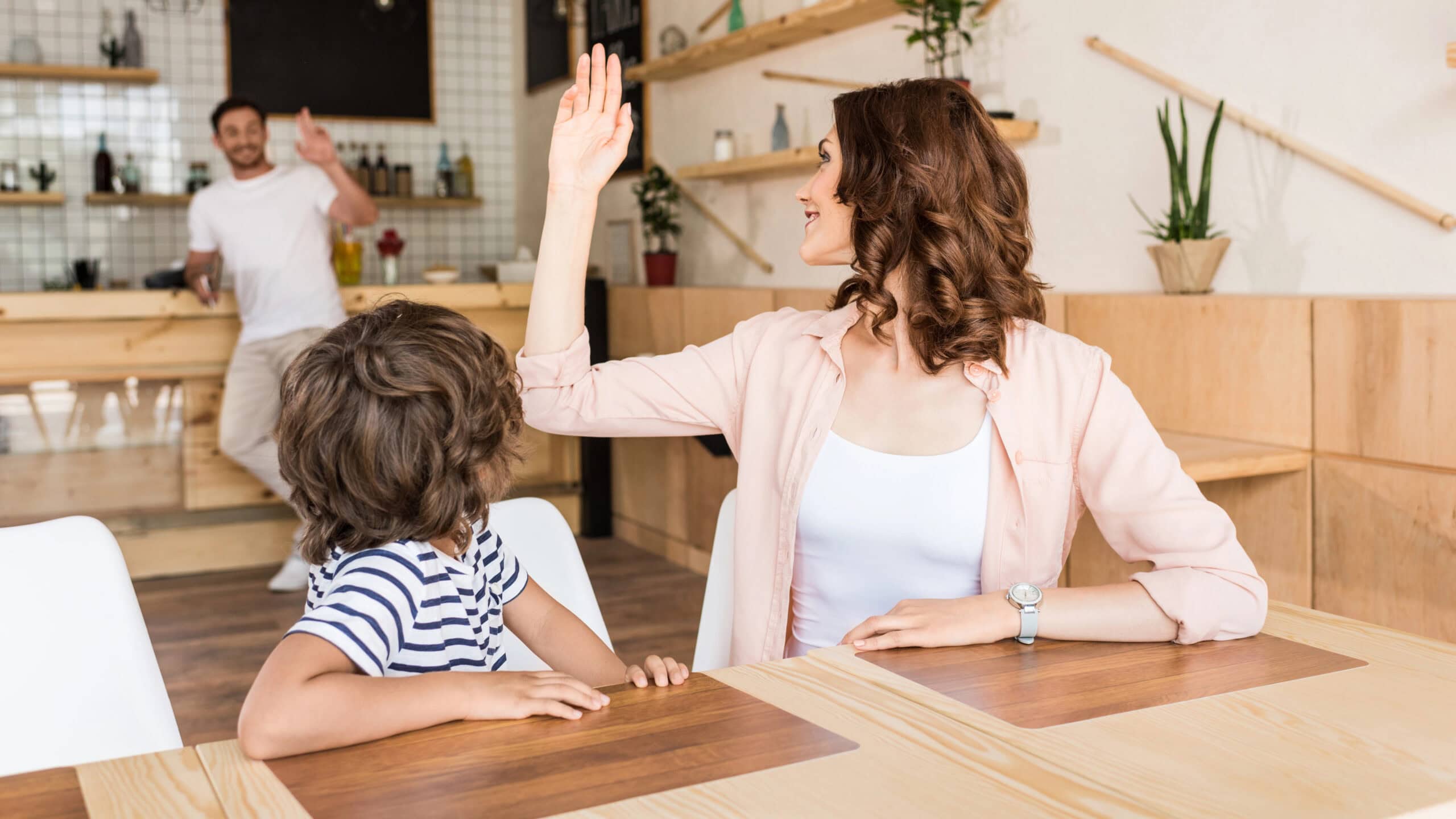 A woman and a child sit at a table in a cafe as the woman raises her hand to wave at a man entering from the background.