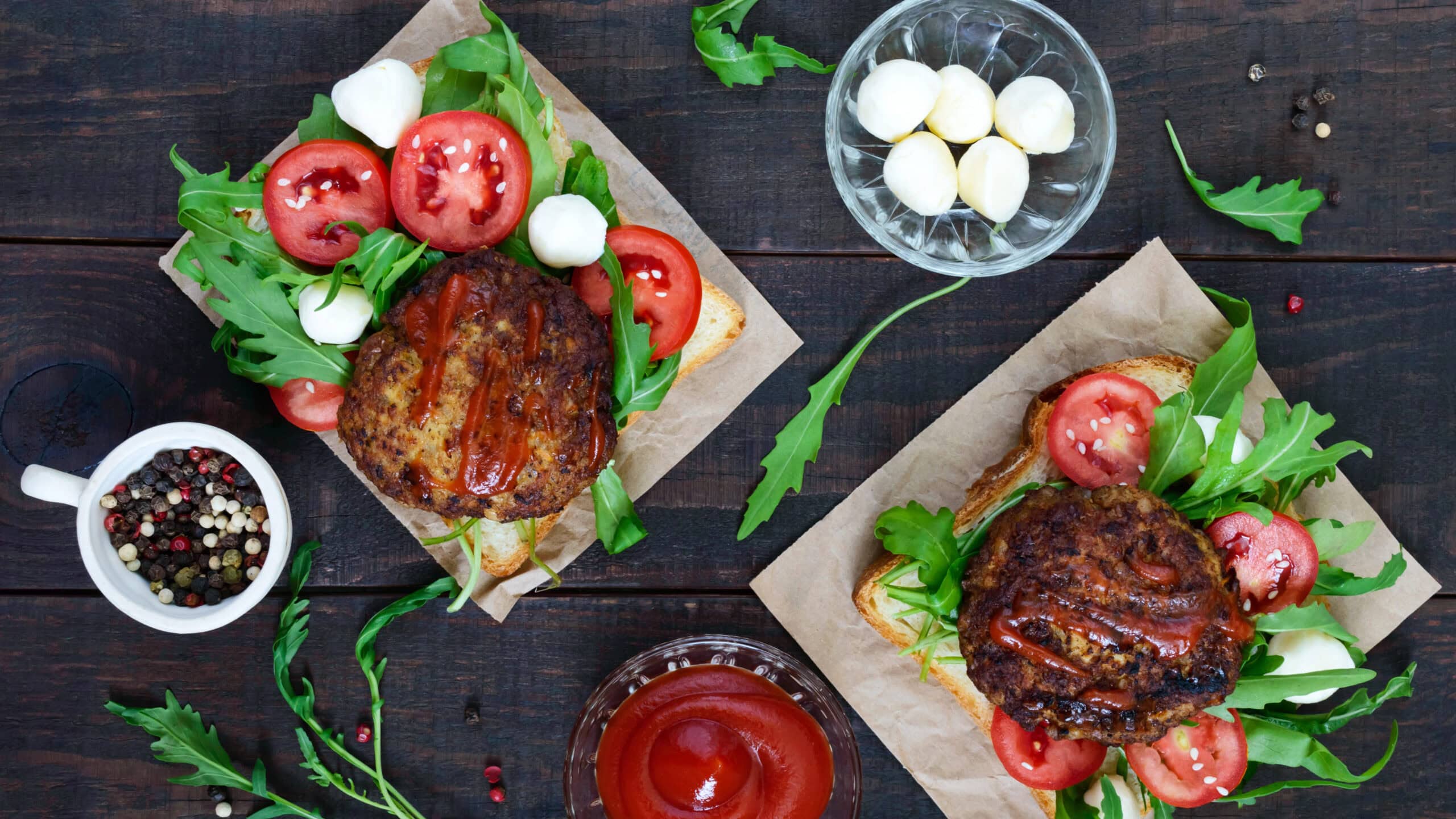 Two burgers with lettuce, tomatoes, mozzarella, and sauce on wooden table. Side dishes include a bowl of mozzarella balls, mixed peppercorns, and a bowl of ketchup.