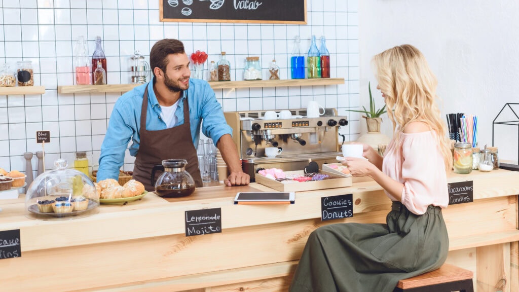 A barista stands behind a café counter talking to a woman holding a cup; pastries, a coffee machine, and a menu board are visible.