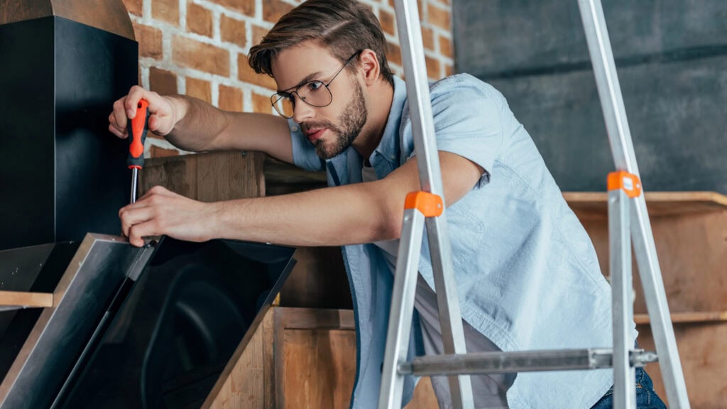 A man wearing glasses uses a screwdriver to repair or assemble a black metal appliance, with a ladder in the foreground and a brick wall in the background.