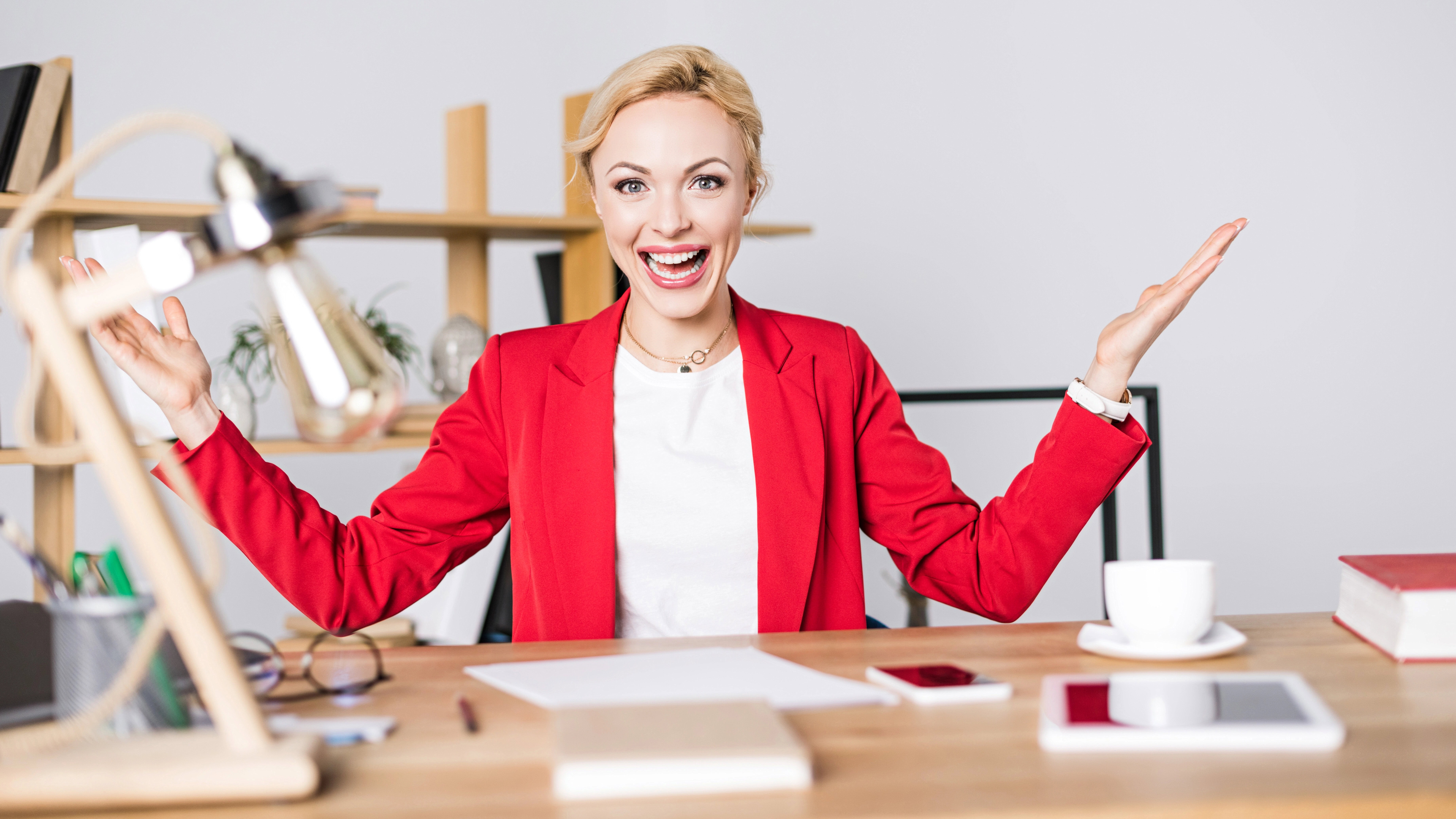 Person in a red blazer sits at a desk with open arms and a cheerful expression, surrounded by office items like a lamp, books, a smartphone, and a cup.
