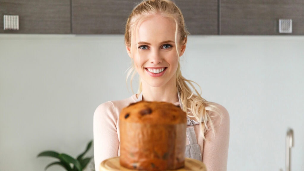 A person in an apron holds a freshly baked panettone on a wooden plate, smiling. A plant is in the background on a kitchen counter.