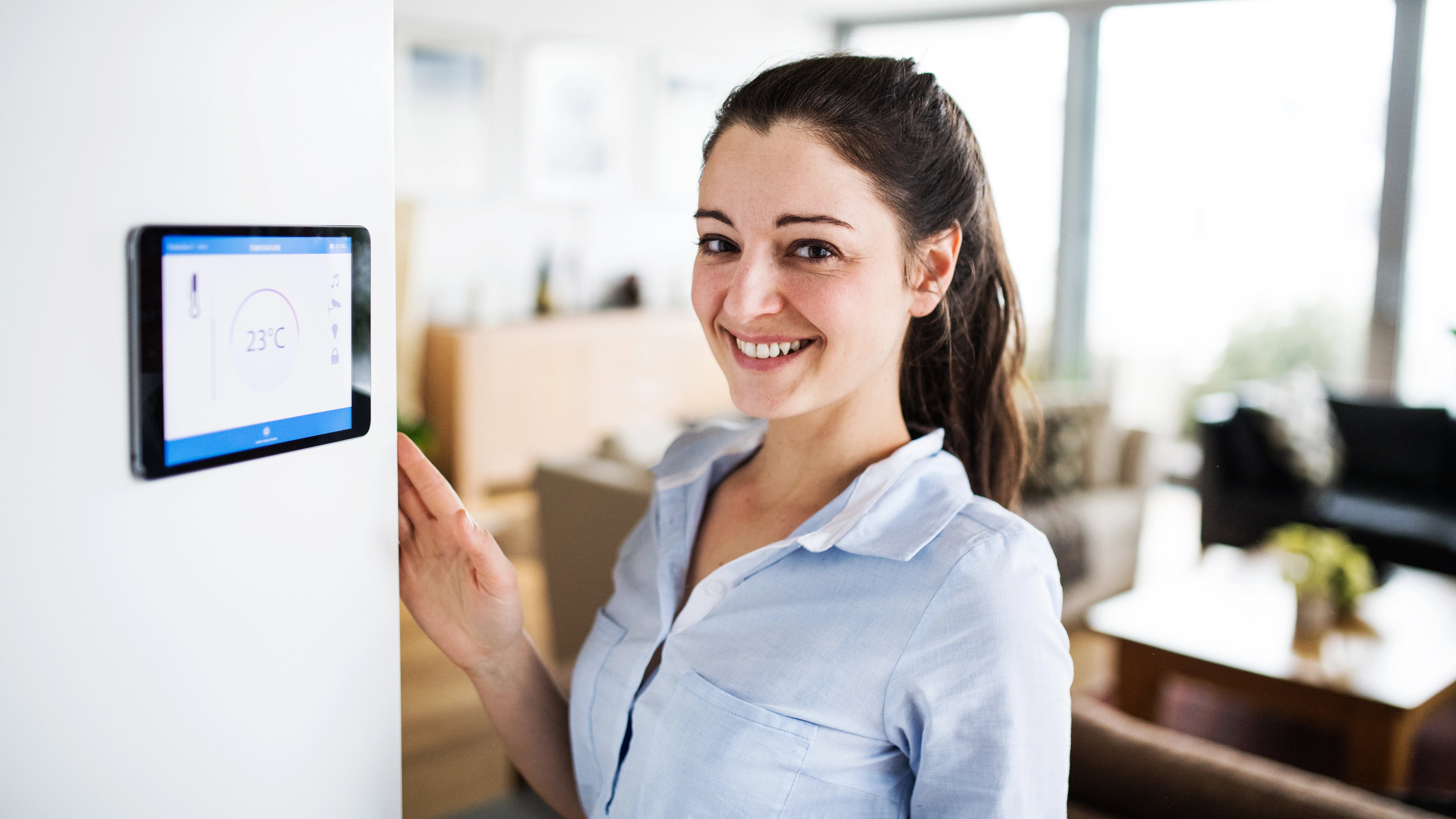 A woman smiles while adjusting a smart home thermostat mounted on a wall.