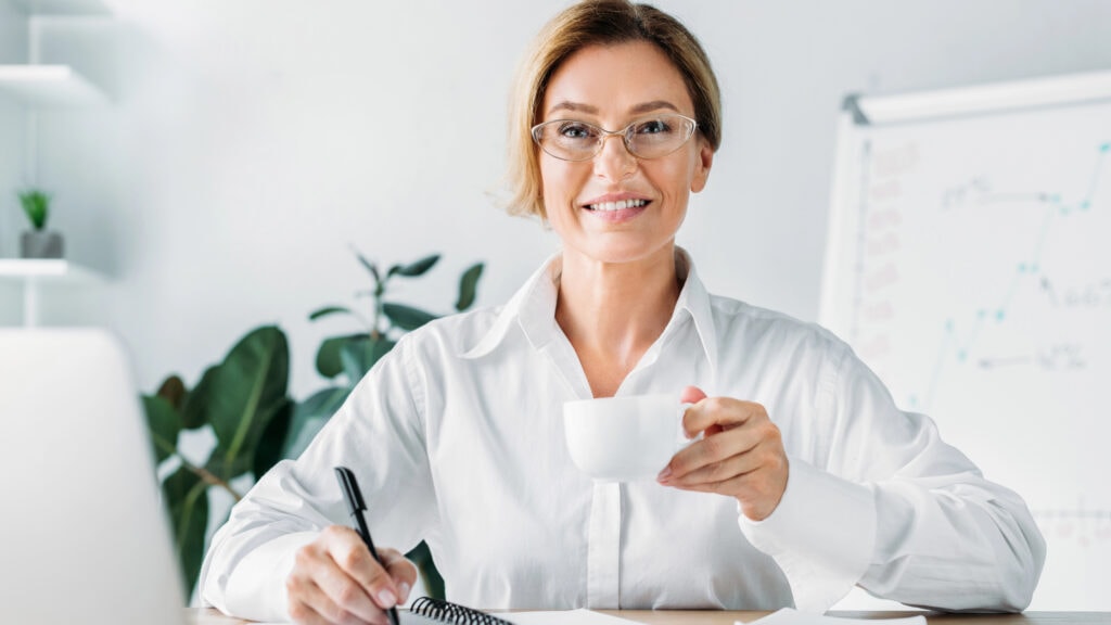 A woman in glasses sits at a desk, holding a white cup, writing in a notebook, with a laptop and a chart in the background.