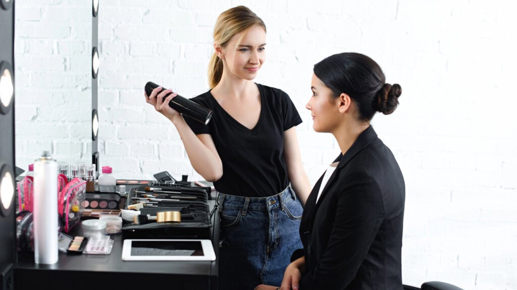 Makeup artist in black t-shirt holds a spray bottle, talking to a seated woman in a black jacket. Various cosmetics and brushes are arranged on the nearby table.