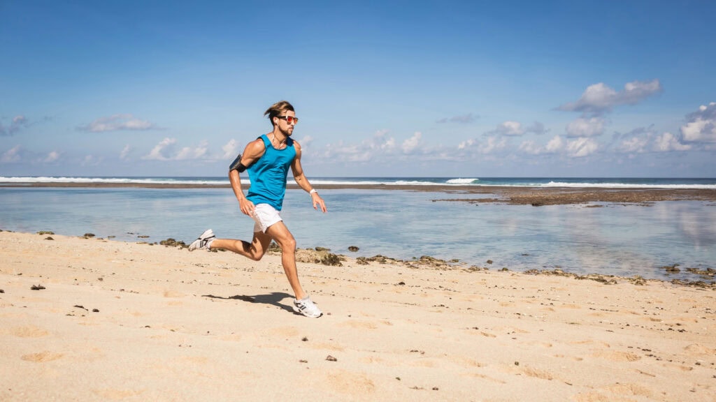 A man in a blue tank top and white shorts runs on a sandy beach near the ocean under a clear sky.