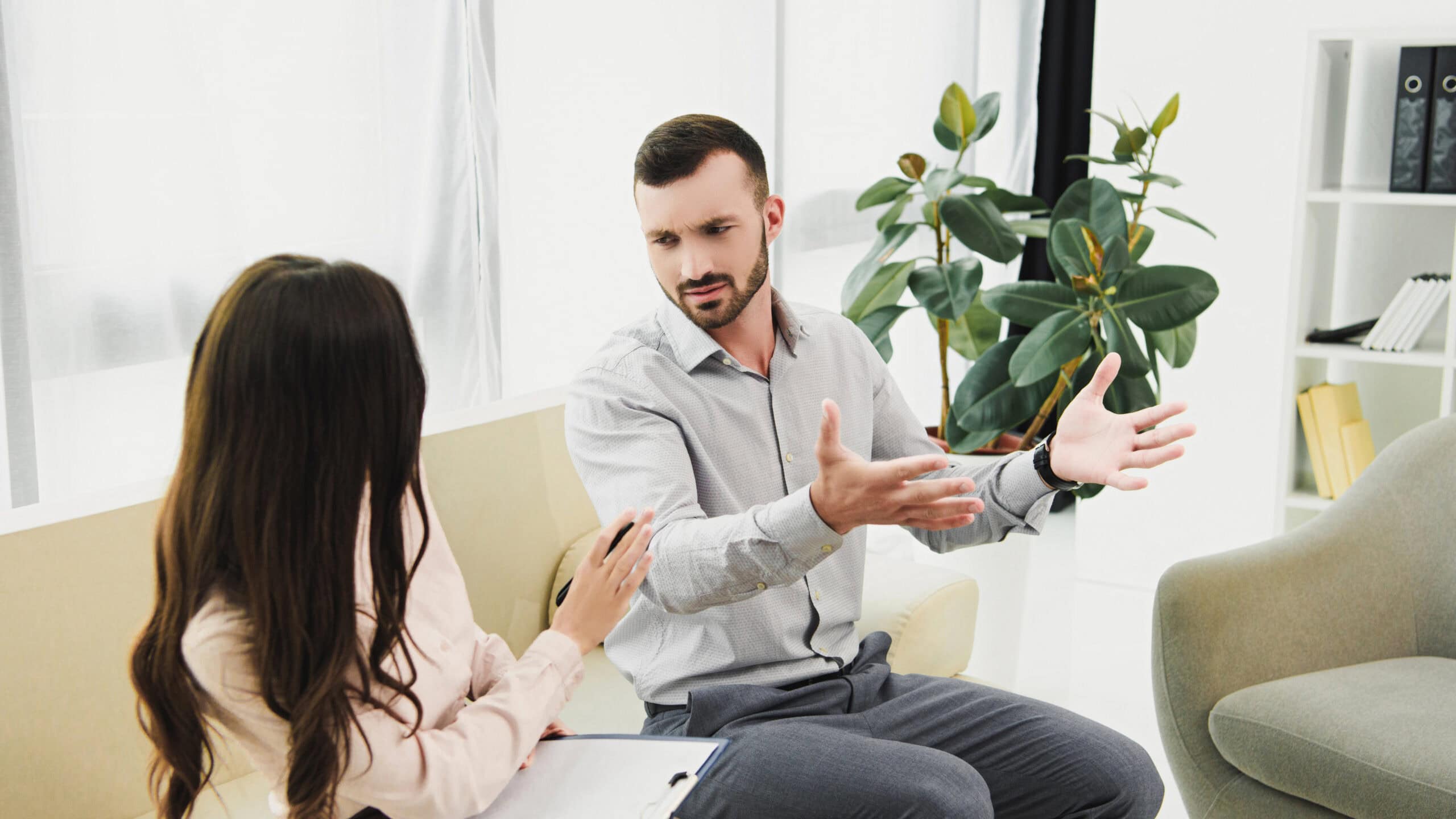 A man and woman are sitting on a couch in an office setting, engaged in a discussion. The man gestures with his hands while the woman holds a clipboard. Plants and books are in the background.