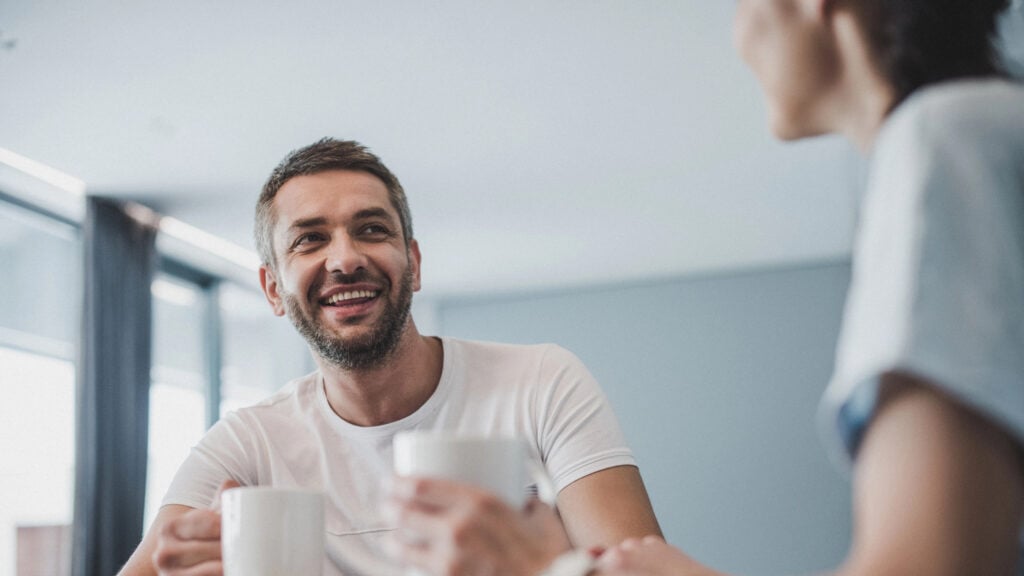 Two people sitting at a table, smiling and holding mugs.