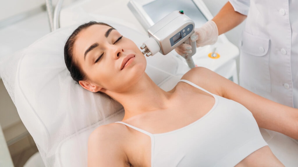 A woman lies on a treatment table with eyes closed while a clinician uses a handheld device on her face. She is wearing a white headband and top.