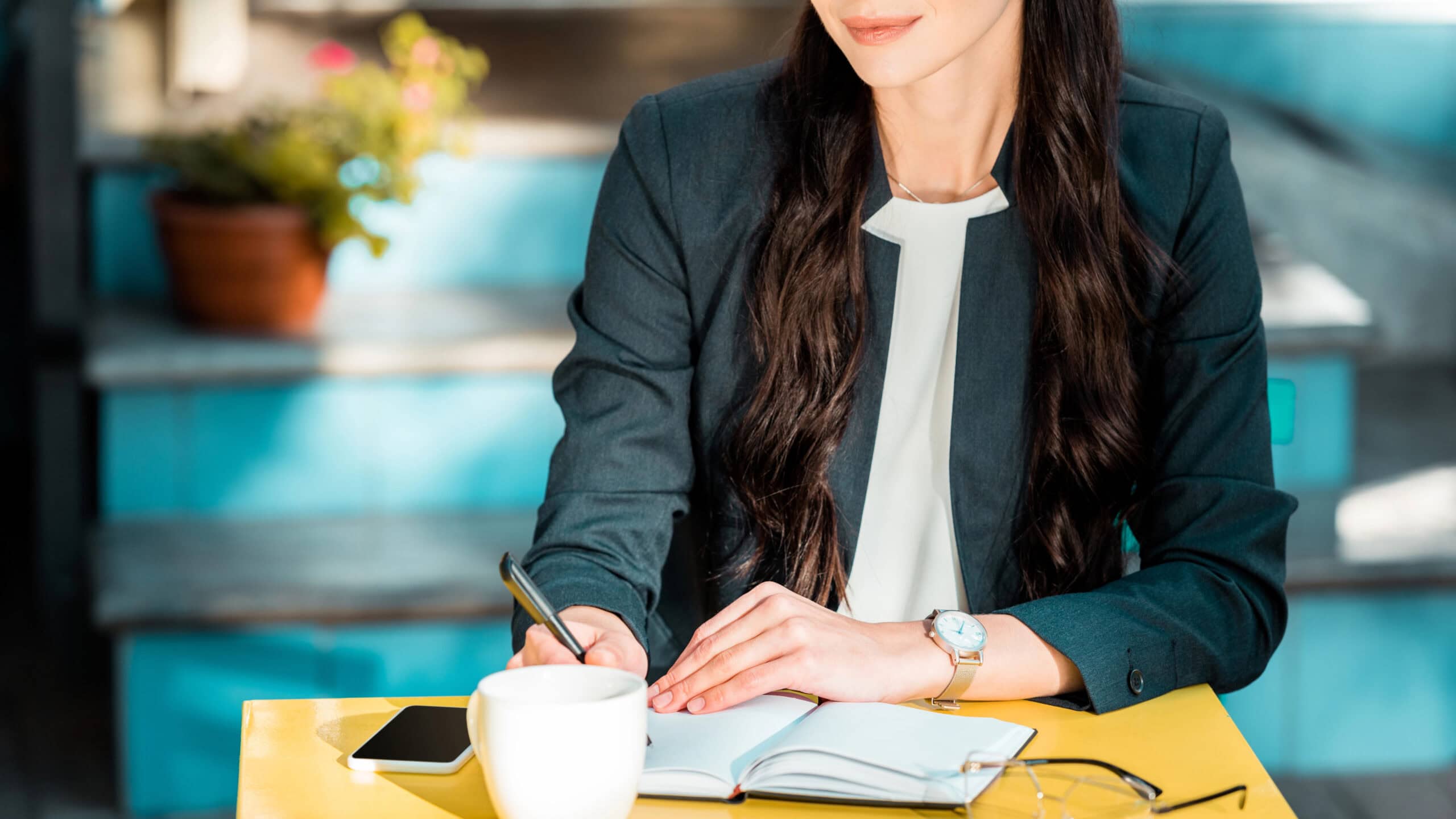 A woman with long hair sits at a yellow table, writing in a notebook. A cup and eyeglasses are nearby. Clipboard in focus, face not shown.