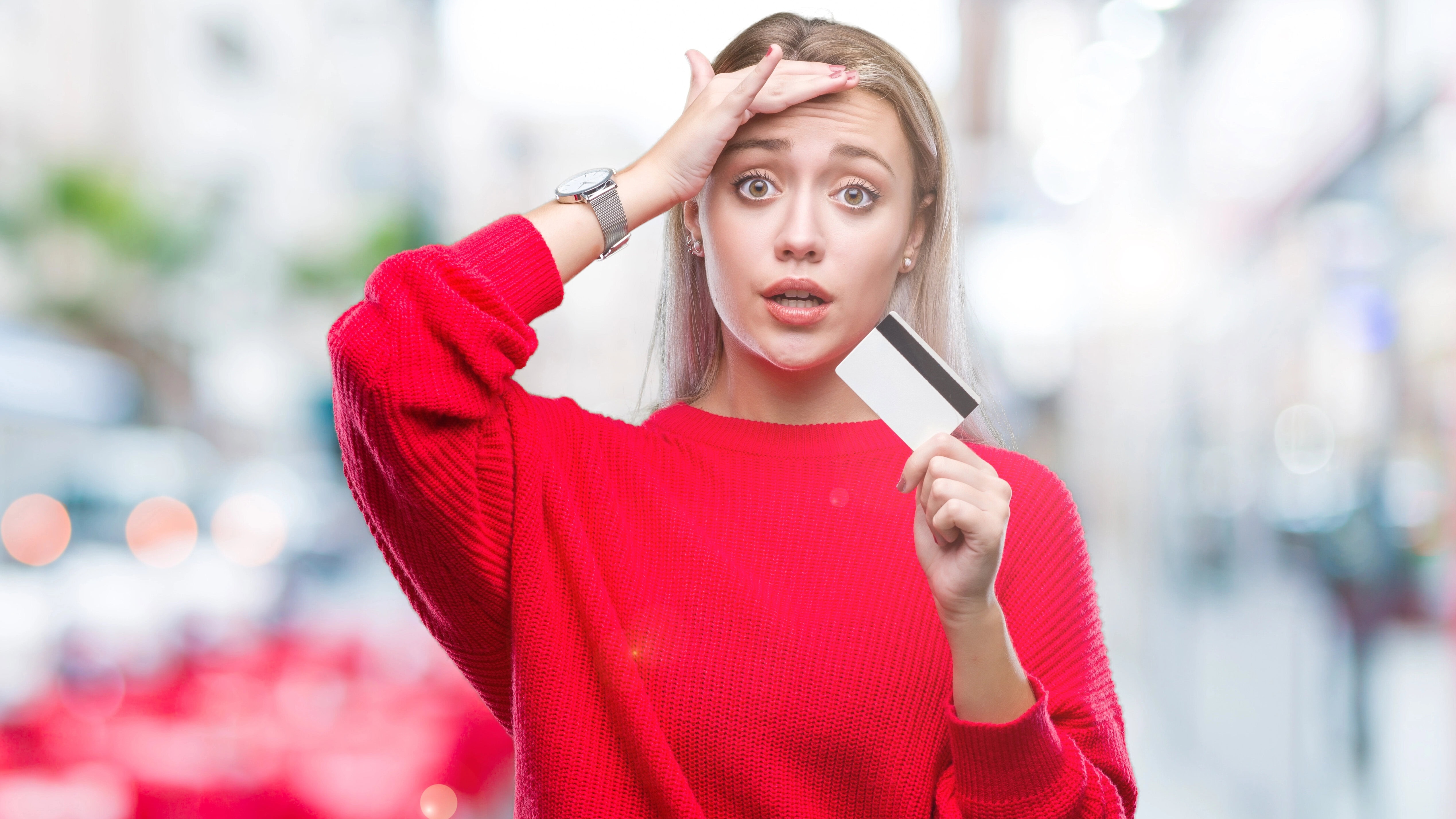Woman in a red sweater holds a credit card, with one hand on her forehead, and an expression of surprise, standing in an outdoor setting with blurred background.