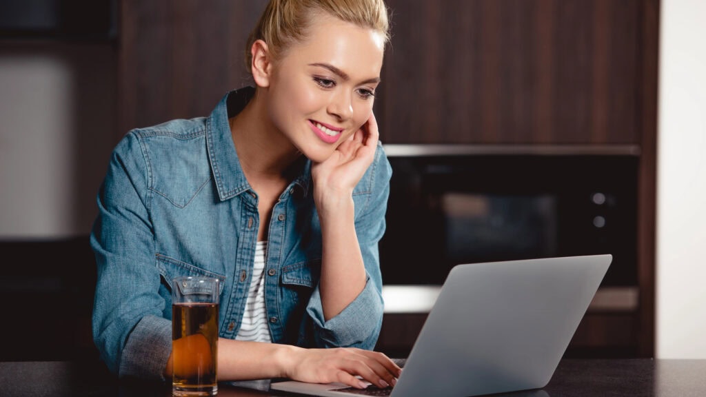 Woman in denim jacket using a laptop at a kitchen counter, with a glass of iced tea nearby, and wood cabinets in the background.