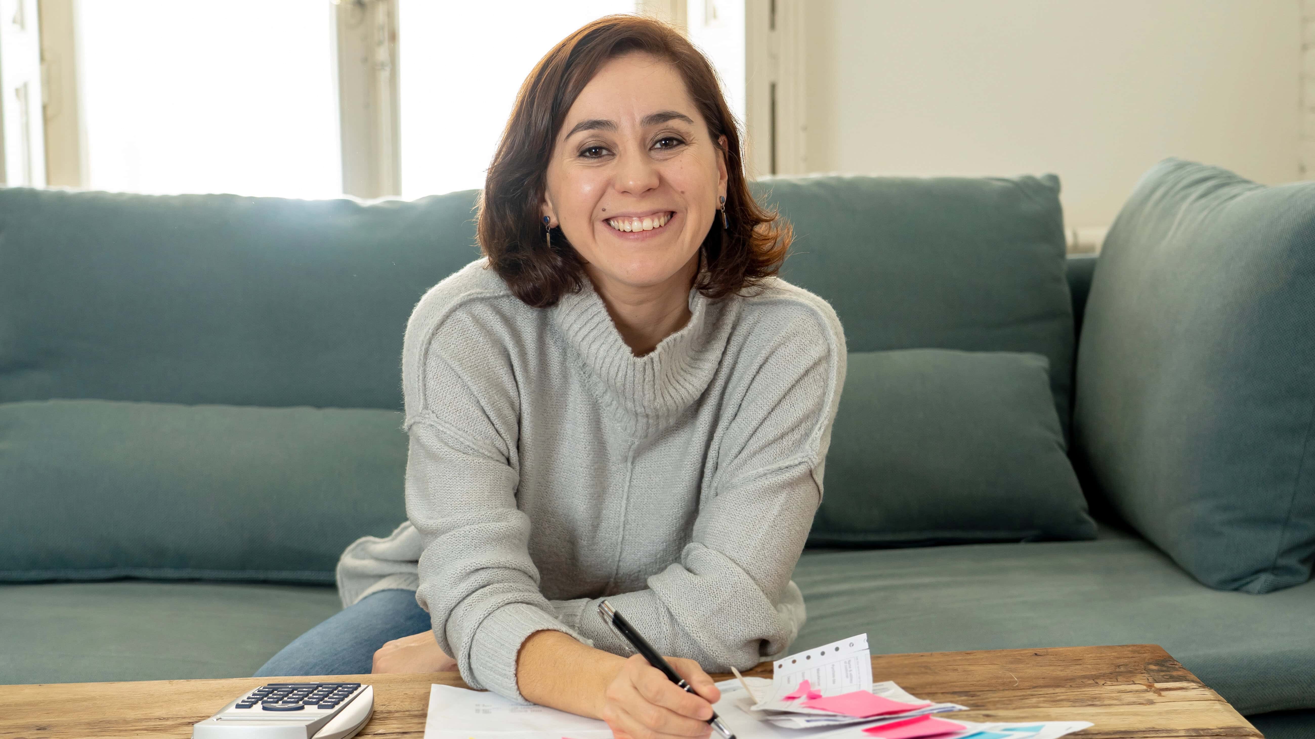 Person smiling, seated on a sofa, writing on papers with a pen. A calculator is nearby on a wooden table. Bright room with natural light from windows in the background.
