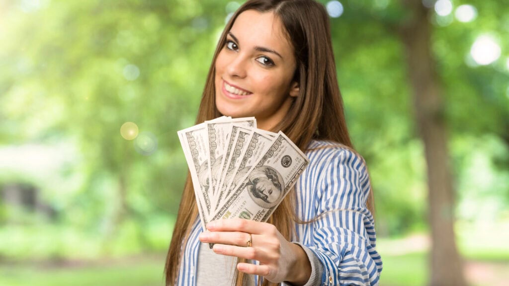 A woman smiles outdoors while holding several hundred-dollar bills fan-like in her hand.