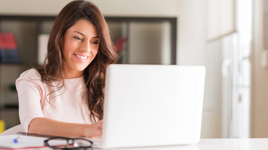 Woman smiling while using a laptop at a desk, with eyeglasses and a notebook nearby.