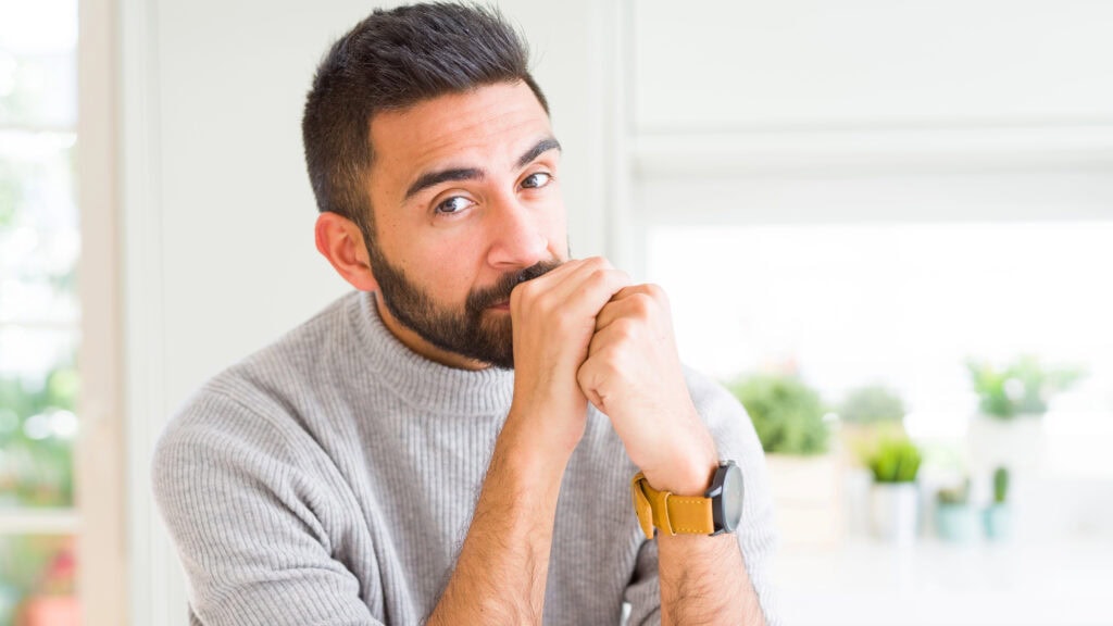 Man with a beard in a gray sweater, sitting indoors with hands near his face, wearing a yellow wristwatch.