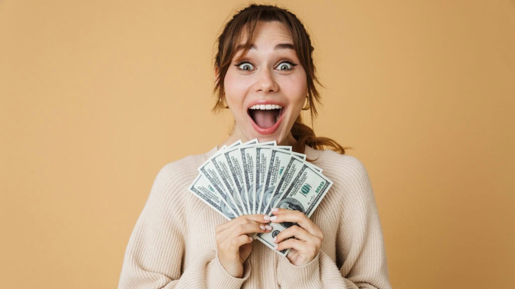 A person with a surprised expression holds several US dollar bills in front of a beige background, seemingly astonished by earnings from underrated side hustles that pay well.