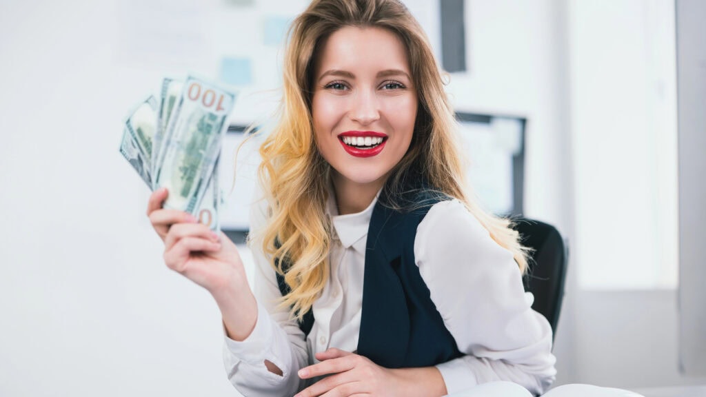 A woman sitting at a desk holds multiple hundred-dollar bills in her hand, smiling. An open book and pen are visible in front of her.