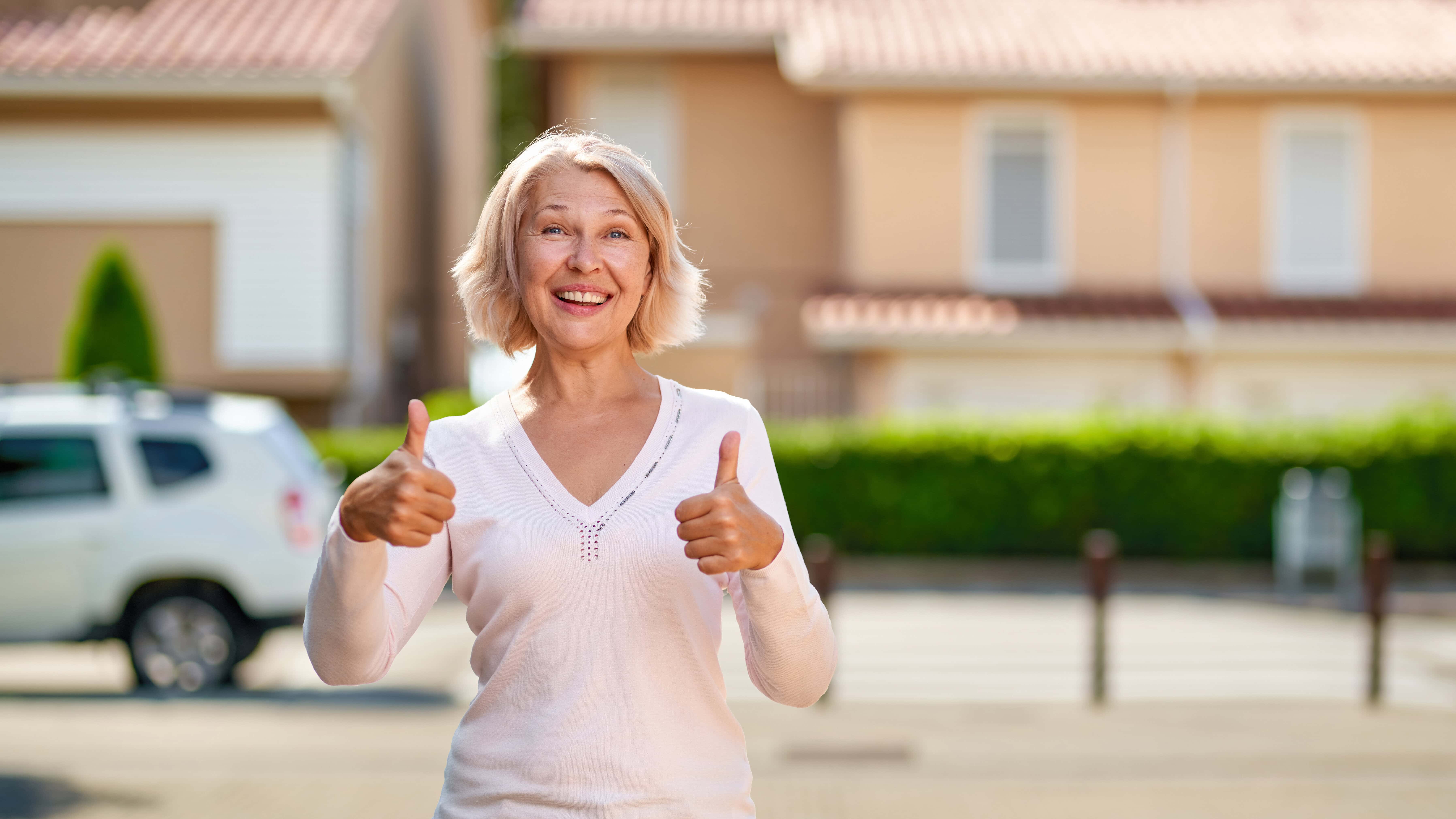 Elderly woman smiling, giving two thumbs up, standing outside with houses and a white car in the background.