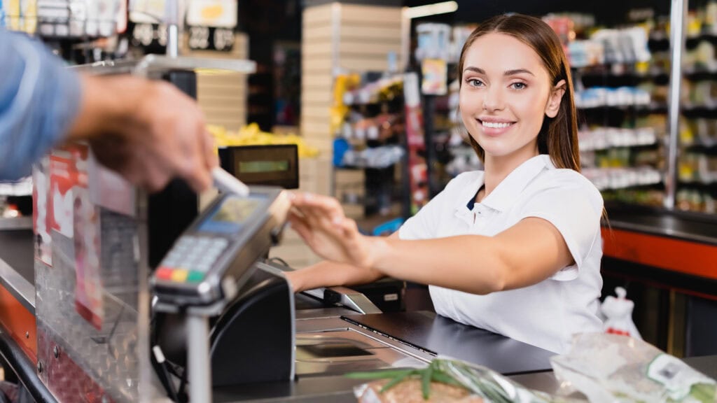 A cashier in a white shirt smiles at the camera while a customer pays with a card at the checkout in a grocery store.