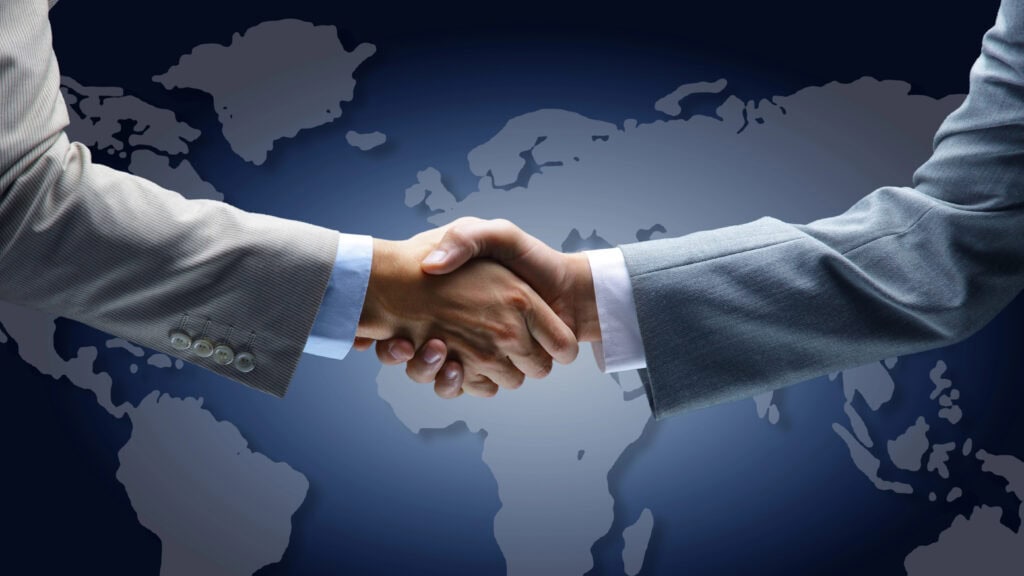Two people in business suits shaking hands in front of a world map, symbolizing a global agreement or partnership.