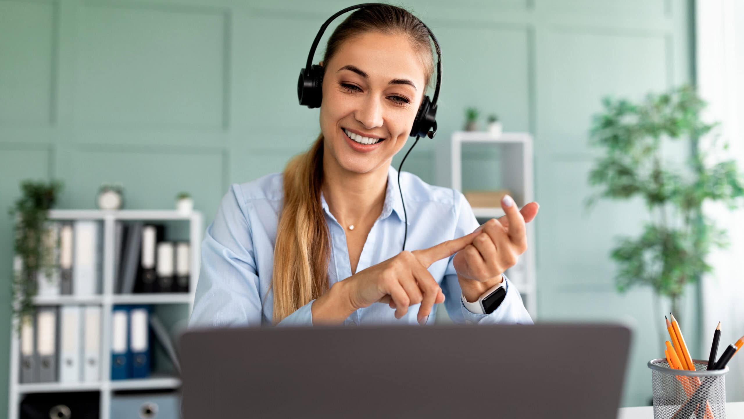 Person wearing headphones, smiling, and pointing at fingers in front of a laptop in a home office with a plant and shelves in the background.