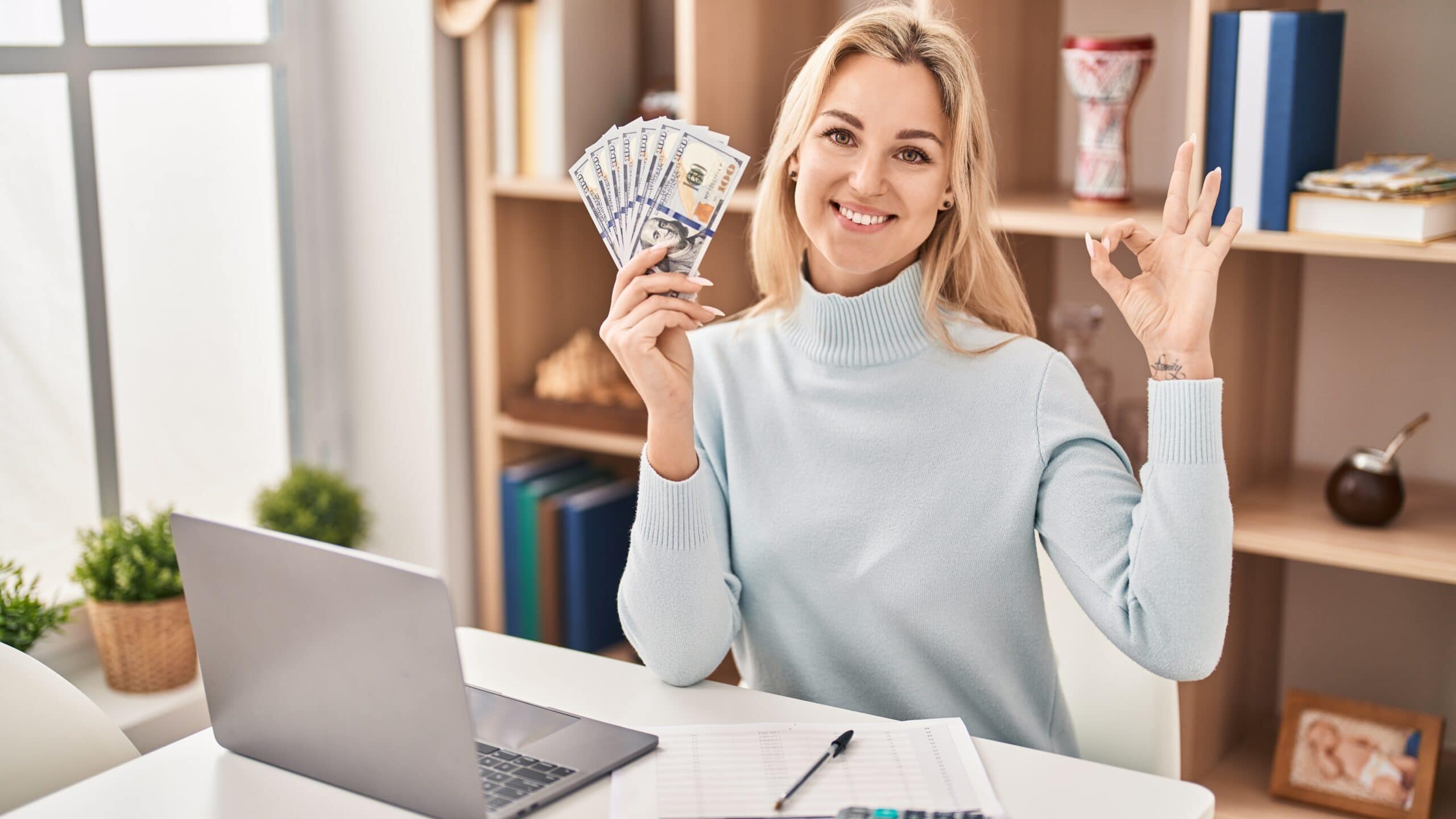 A person sitting at a desk with a laptop and calculator, holding dollar bills in one hand and making an "OK" gesture with the other, smiling. Books and plants are on the shelves behind.