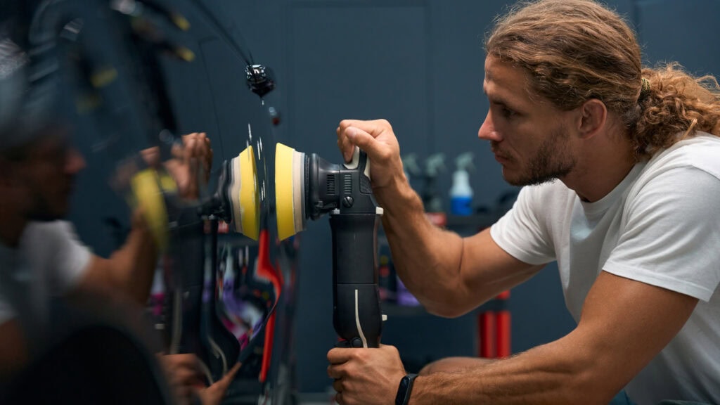 A man in a white t-shirt and shorts is using a polishing tool on a car's surface in a garage-like setting.