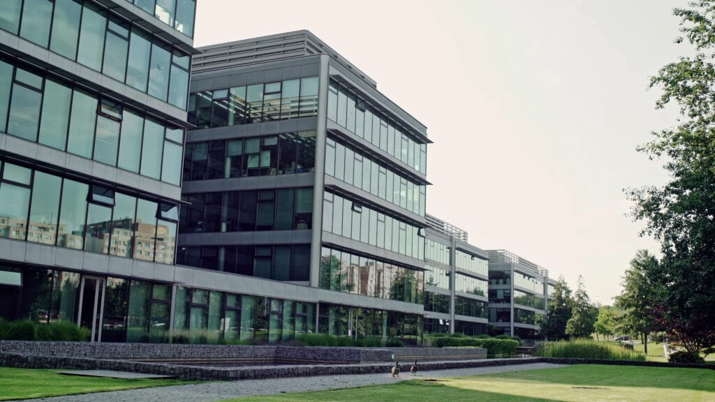 Modern glass office buildings with multiple floors, large windows, and landscaped green space in the foreground on a sunny day.