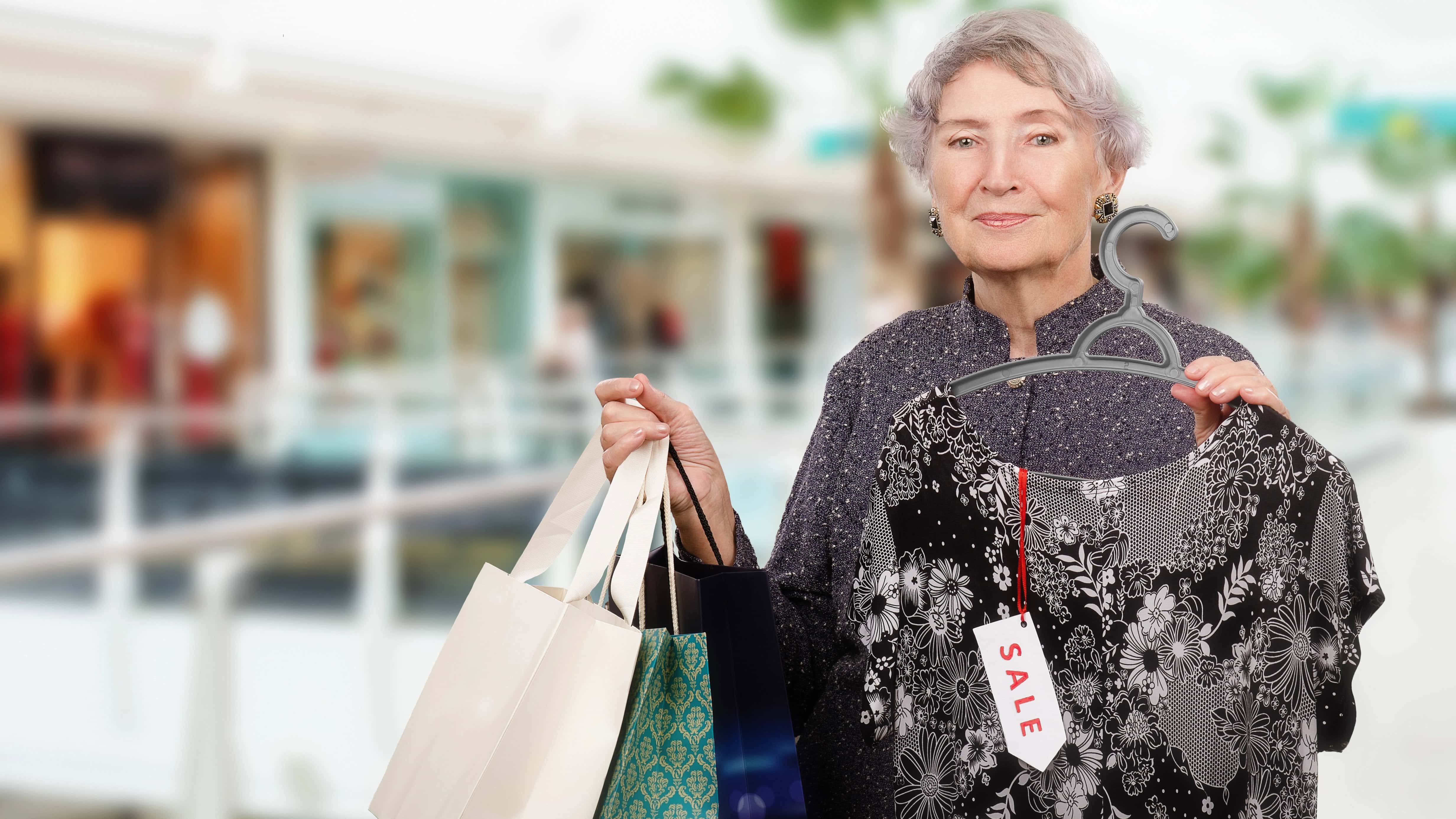 Older woman in a shopping mall holding several shopping bags and a floral dress on a hanger with a sale tag attached.