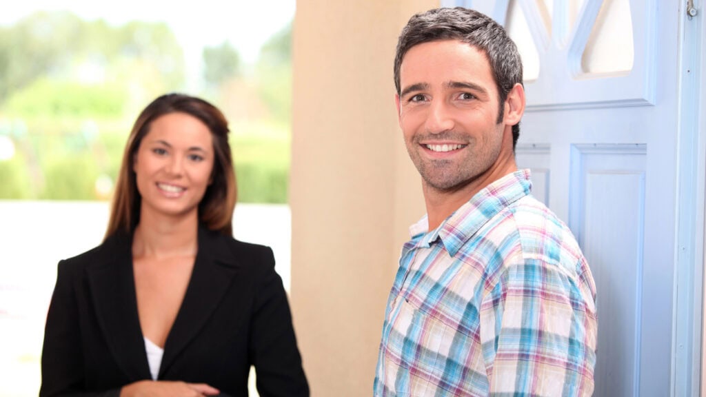 A man in a plaid shirt stands by a door, smiling at the camera, with a woman in a black blazer smiling in the background.