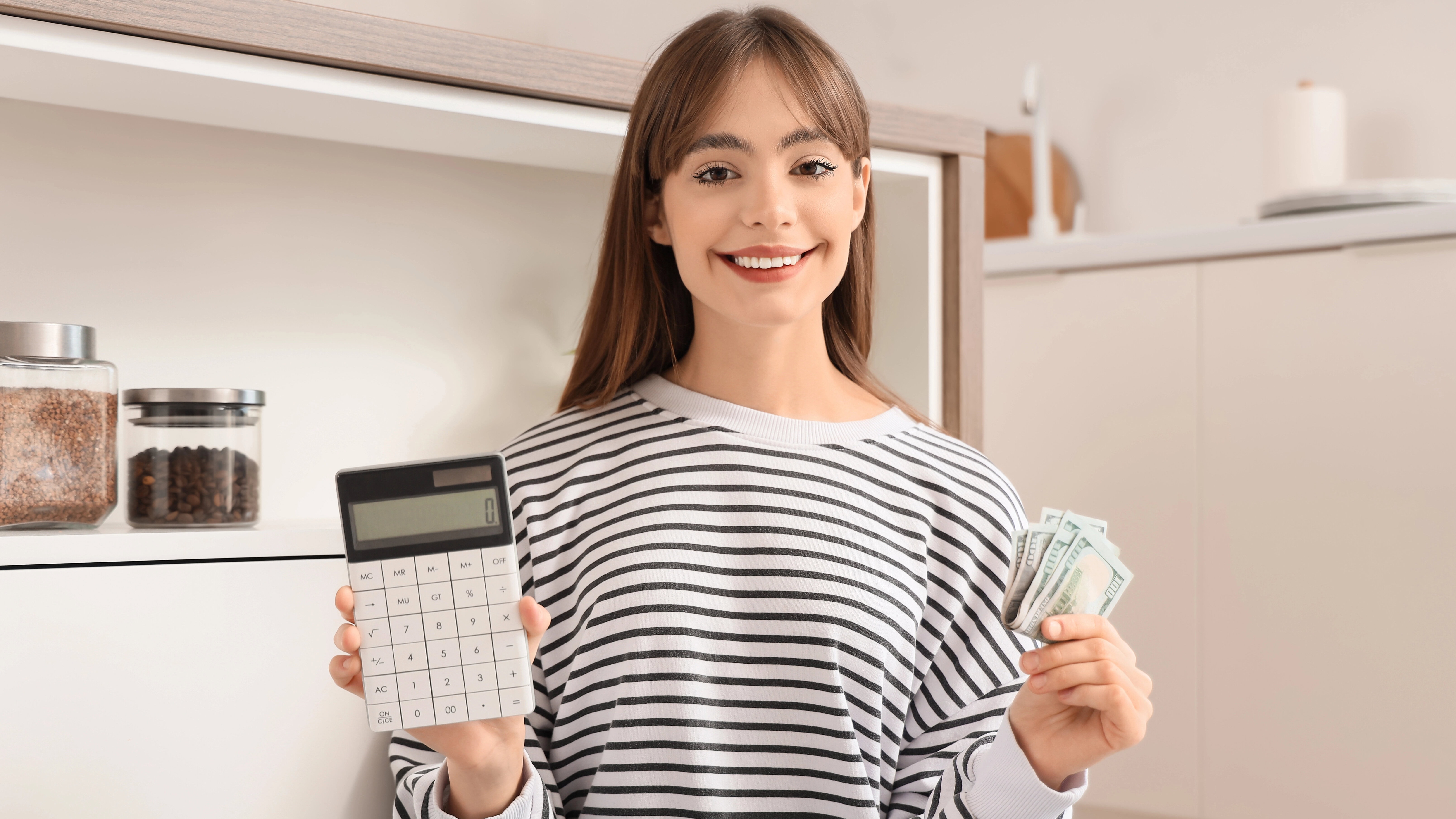 Woman in a striped shirt sitting on the floor, holding a calculator in one hand and dollar bills in the other, smiling at the camera.