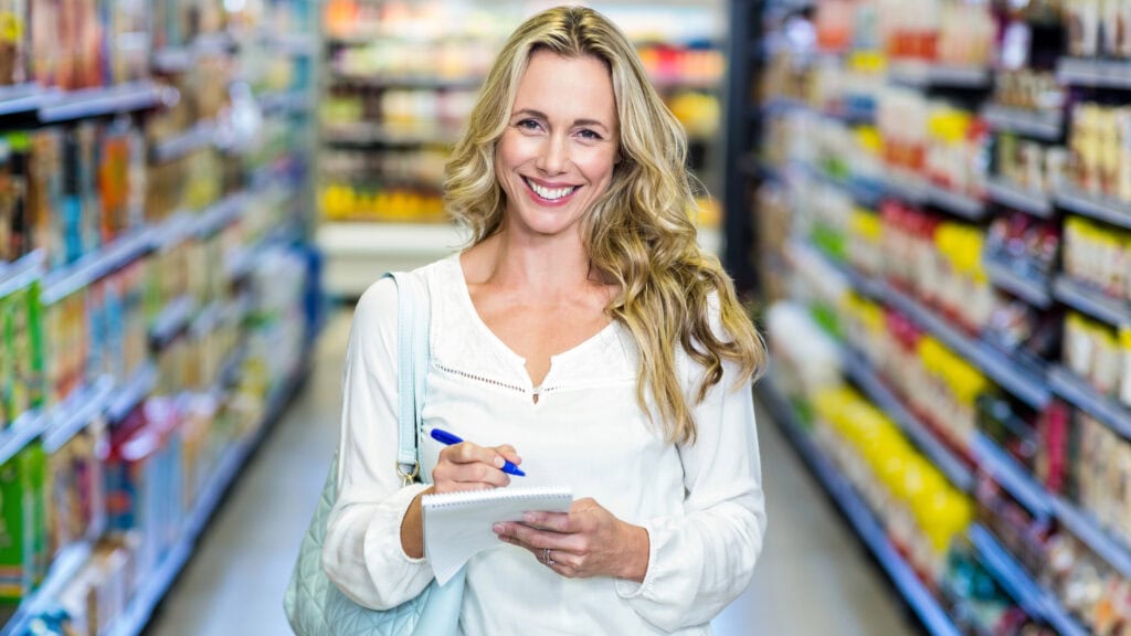 Woman with long blonde hair writes in a notepad while standing in a grocery store aisle filled with various products.
