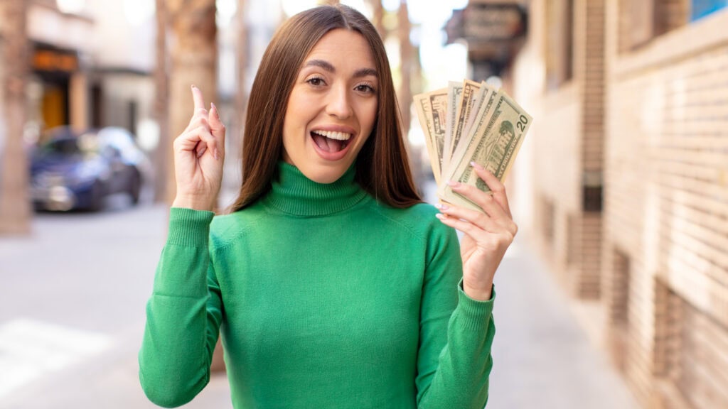 A woman in a green sweater smiles and holds a fan of U.S. dollar bills while gesturing with her other hand on a city street.
