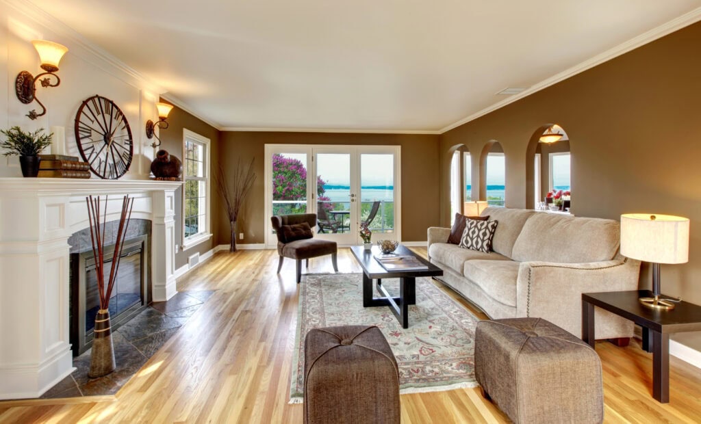 A cozy living room with a beige sofa, coffee table, and fireplace. Large windows and a glass door provide a view of a patio and greenery. Warm lighting and wooden flooring create a welcoming ambiance.