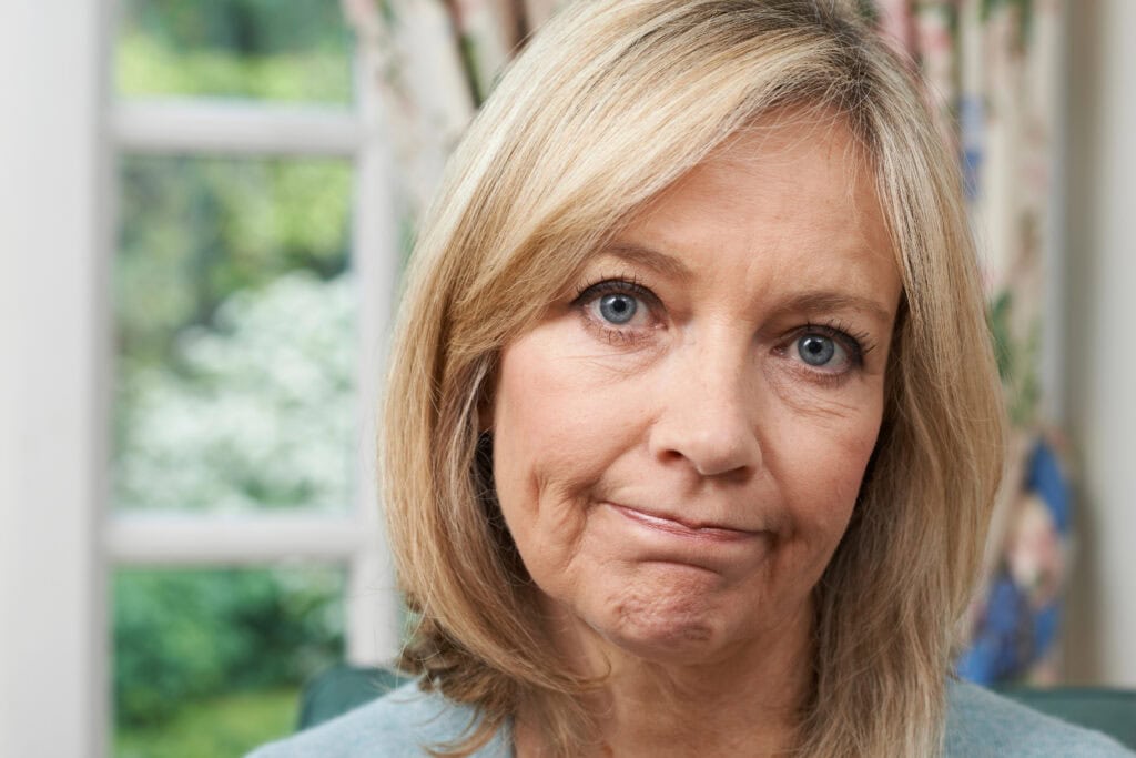 A woman with shoulder-length blonde hair looks directly at the camera with a neutral expression, sitting indoors by a window.