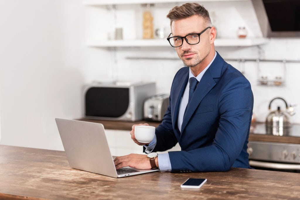 Businessman in eyeglasses holding cup of coffee and looking at camera while using laptop at home