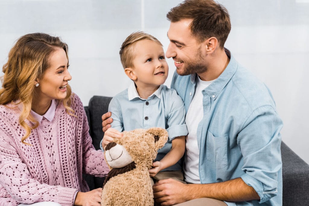 Cheerful parents sitting on sofa and embracing cute son holding teddy bear in apartment