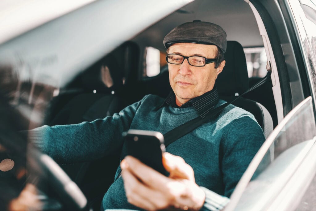 Senior man with cap on head and eyeglasses sitting in car and using smart phone.