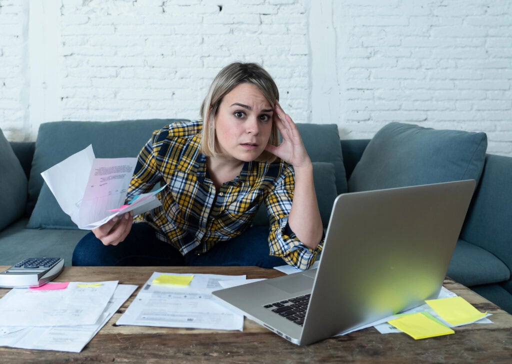 A woman looks stressed while reviewing documents at a table, with a laptop, calculator, and papers around her.
