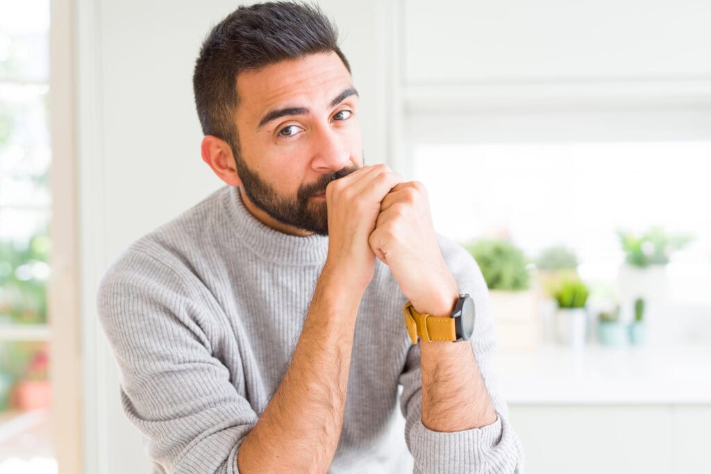 Man with a beard in a gray sweater, sitting indoors with hands near his face, wearing a yellow wristwatch.