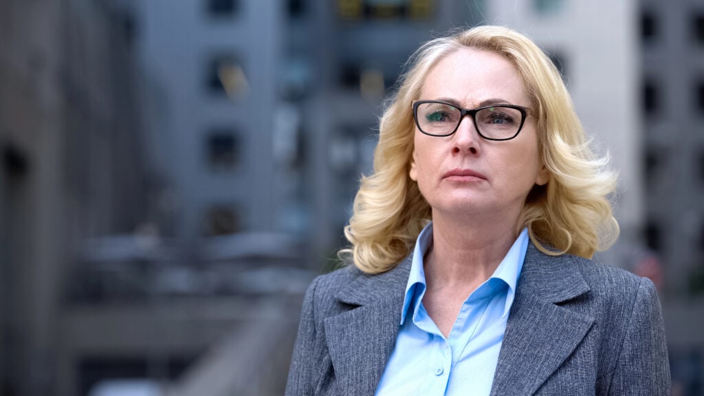 Woman with blonde hair and glasses in a gray blazer stands outdoors in an urban setting.