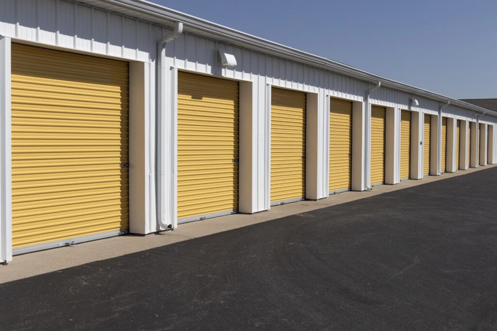 A row of storage units with closed yellow garage doors, set against a white building, beneath a clear blue sky.
