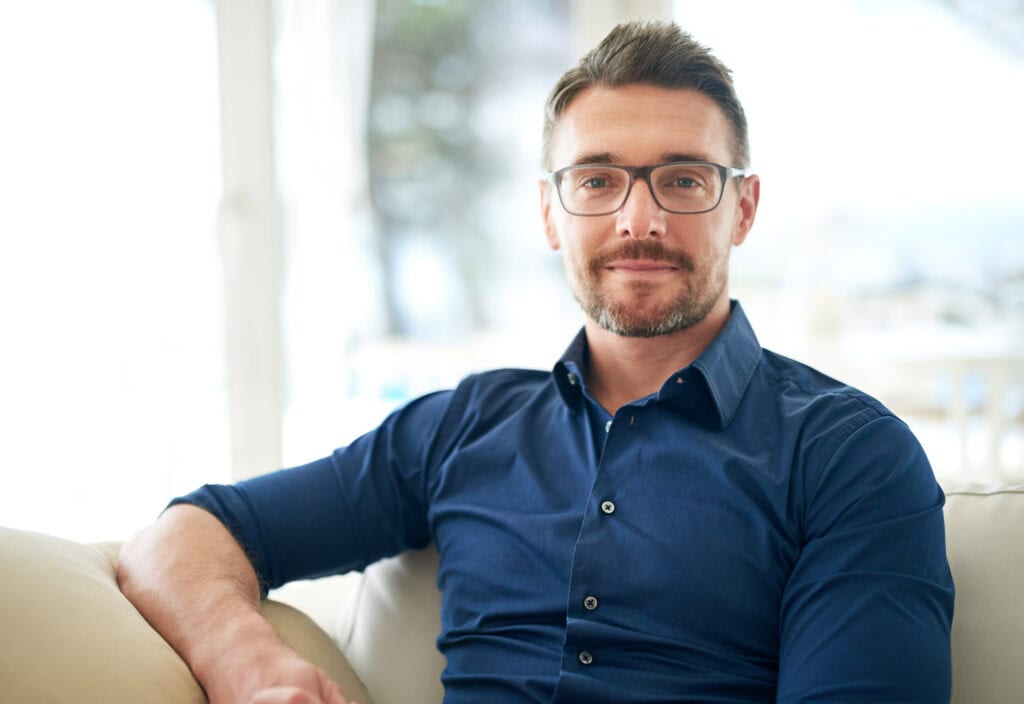 A man with glasses and a beard sits on a couch, wearing a blue shirt. The background is softly blurred.