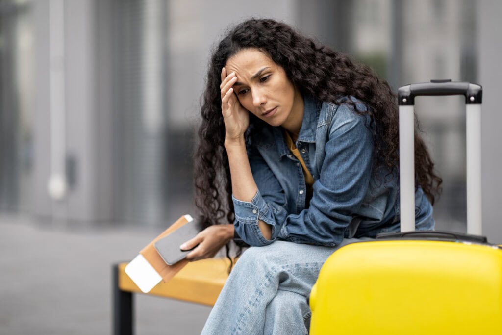 A woman with long curly hair, wearing a denim jacket and jeans, sits on a bench holding a passport and ticket. She appears tired or stressed, with a yellow suitcase beside her.
